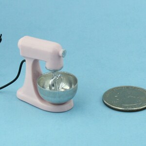 以下が含まれることがあります： 銀色のボウルと泡立て器アタッチメントが付いた、ミニチュアのピンク色のスタンドミキサー。黒いコードがミキサーから伸びています。サイズの比較のために、ミキサーの隣にコインがあります。ミキサーはおもちゃまたは装飾品です。