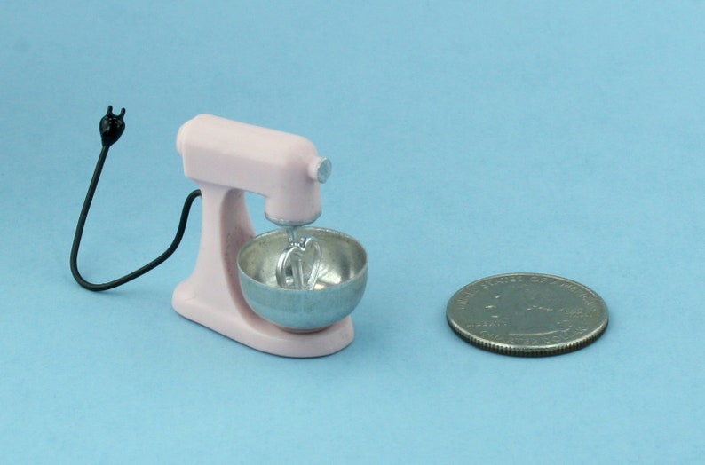 以下が含まれることがあります： 銀色のボウルと泡立て器アタッチメントが付いた、ミニチュアのピンク色のスタンドミキサー。黒いコードがミキサーから伸びています。サイズの比較のために、ミキサーの隣にコインがあります。ミキサーはおもちゃまたは装飾品です。