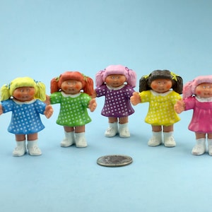 May include: Five small doll figurines with different colored dresses and hair, including blue, green, purple, yellow, and pink. Each doll has white boots and is standing on a light blue surface. A coin is in front of the dolls.