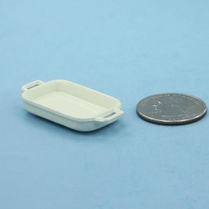 May include: A small, off-white rectangular baking dish with handles, next to a quarter coin on a light blue surface. The dish is empty and appears to be made of ceramic or porcelain. The coin provides a size reference.