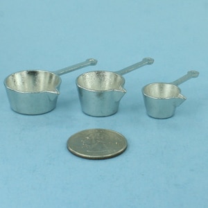 May include: Three small, silver-colored metal saucepans with handles, each with a pouring spout. A US quarter is next to the saucepans for size comparison. The saucepans are on a light blue surface.