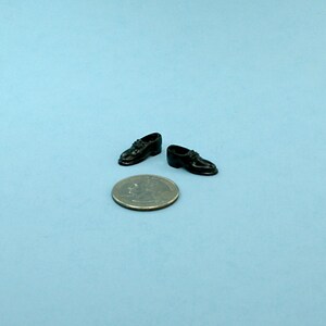 May include: Miniature black dress shoes with laces are displayed next to a quarter coin on a light blue surface. The shoes are glossy and appear to be made of a hard material. The shoes are a small size.