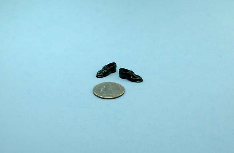 May include: Miniature black dress shoes with laces are displayed next to a quarter coin on a light blue surface. The shoes are glossy and appear to be made of a hard material. The shoes are a small size.