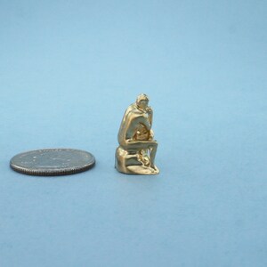 May include: A gold-colored miniature sculpture of a seated figure, reminiscent of the "Thinker" statue, is shown next to a US quarter for scale.