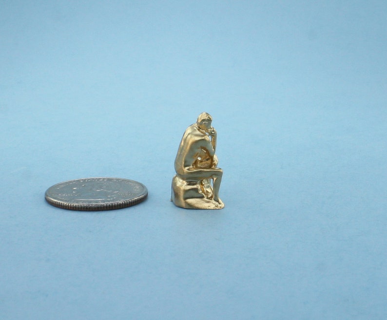 May include: A gold-colored miniature sculpture of a seated figure, reminiscent of the "Thinker" statue, is shown next to a US quarter for scale.