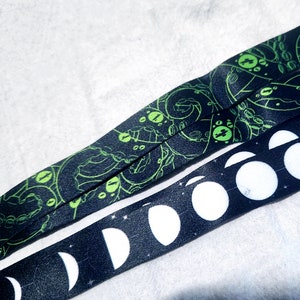 Op de afbeelding: Twee zwarte en groene gepatroneerde lanyards. De ene lanyard heeft een herhalend patroon van een groen wezen met grote ogen. De andere lanyard heeft een herhalend patroon van witte halve manen op een zwarte achtergrond.