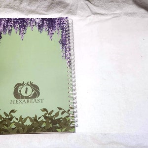 May include: A green notebook with a spiral binding. The cover features a purple floral design, a stylized eye logo, and the text "HEXABEAST".