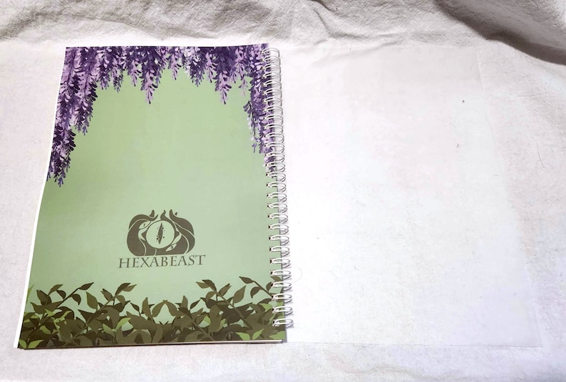 May include: A green notebook with a spiral binding. The cover features a purple floral design, a stylized eye logo, and the text "HEXABEAST".