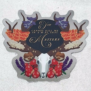 Könnte beinhalten: Silberner Aufkleber mit einem Design aus Hirschschädel und Pilzen. Der Aufkleber enthält den Text "You cannot kill me in a way that Matters". Das Design umfasst rote, violette und orangefarbene Pilze und Lavendel.