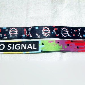 May include: Two black and white patterned wristbands. The top band features a repeating eye design with red, blue, and white pixelated details. The bottom band has the text "NO SIGNAL" in white letters on a black background with a colourful pixelated design.