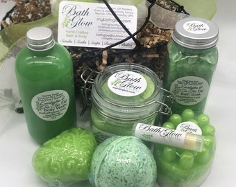 May include: A green and white gift basket filled with bath and body products. The basket includes a green body wash, a jar of bath salts, a bath bomb, a bar of soap, and a lip balm. The products are all labeled with the brand name "Bath Glow".