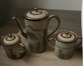 May include: A set of three white porcelain teacups with gold accents and floral designs. The teapot has a gold lid and a handle. The cups have a gold rim and a handle.