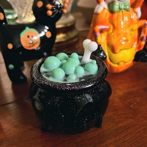 May include: A black ceramic cauldron with green bubbles and a bone, with a black cat and a ghost figurine in the background. The cauldron has a glittery rim. The cat has orange spots and a pumpkin on its side. The ghost is orange and white.