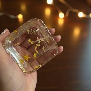 May include: A clear, square resin trinket dish filled with gold glitter and star-shaped confetti. The dish is held in a hand, with a blurred background of warm, glowing lights. The dish is a decorative item.