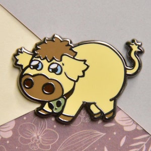 May include: A yellow enamel pin of a cartoon animal with brown fur, brown eyes, and a brown nose. The animal is wearing a green and black collar.