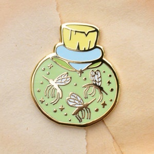 May include: A green enamel pin shaped like a glass jar with a yellow lid and three white mosquitos inside. The jar has a light blue band around the top and is decorated with gold glitter.