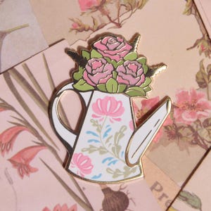 May include: A white and gold enamel pin in the shape of a watering can, decorated with pink floral patterns. The watering can is topped with three pink roses and green leaves. The pin is set against a backdrop of vintage floral ephemera.