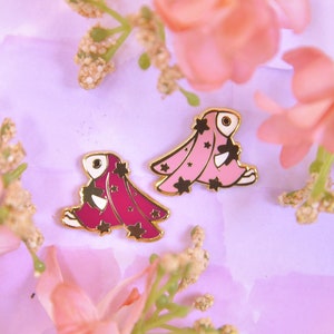 Puede incluir: Dos pines de esmalte dorado con un animal de dibujos animados que lleva una bata rosa y morada con estrellas. Los pines están sobre un fondo morado claro con flores rosas.