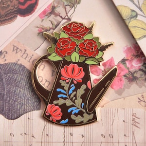 Könnte beinhalten: Ein dekorativer Emaille-Anstecker in Form einer braunen Gießkanne, verziert mit roten Rosen und floralen Designs. Der Anstecker hat eine goldene Umrandung und befindet sich vor einem Hintergrund aus Vintage-Papier und botanischen Illustrationen.