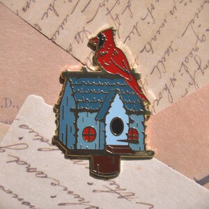 Könnte beinhalten: Detaillierte Emaille-Anstecknadel mit einem roten Kardinal, der auf einem blauen Vogelhaus sitzt. Das Vogelhaus hat ein hellblaues Dach und einen dunkelblauen Körper mit goldenen Verzierungen. Die Nadel befindet sich vor einem Hintergrund aus Vintage-Papier.