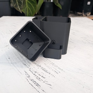 May include: A black, square-shaped plant pot with drainage holes, next to a matching rectangular planter with multiple compartments. The items are made of a matte material and are designed for indoor gardening. The background features other potted plants.