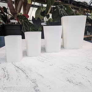 May include: Four white, geometric-shaped plant pots of varying sizes are arranged on a white, weathered wooden surface. The pots are empty, with a clean, modern aesthetic. The background features lush green plants in a greenhouse setting.