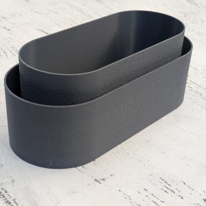 May include: Two nested oval-shaped containers in a dark charcoal gray color. The containers feature a textured exterior surface and smooth interior walls, with an open top design and a continuous, rounded rim that forms a simple, elongated shape.