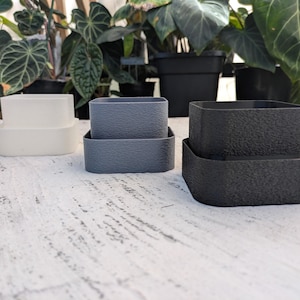 May include: Three sets of two-piece square containers in white, gray, and black. Each set consists of a smaller inner square container nested inside a slightly larger outer square container. The gray and black containers feature a textured surface.