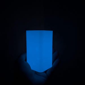 May include: A rectangular object emitting a bright, solid blue glow against a dark background. The object has a smooth, flat surface with straight vertical edges and a slightly rounded bottom edge, creating a uniform light source in the darkness.