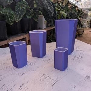 Puede incluir: Un conjunto de tres macetas hexagonales de color morado con bordes interiores blancos. Las macetas varían en altura y anchura, presentando un diseño geométrico liso con lados verticales planos. Cada recipiente está abierto.