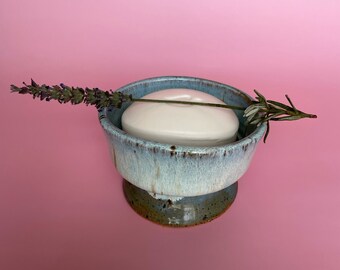 May include: A handmade ceramic soap dish with a light blue glaze, holding a white bar of soap. A sprig of lavender rests across the dish. The dish has a pedestal base and is set against a pink background.
