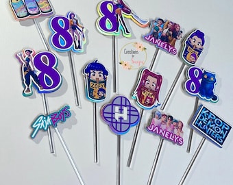May include: A collection of colorful cake toppers on silver sticks. The toppers feature various designs, including the number 8, cartoon characters, and text such as "Six Boys," "Janelys," and "K-Pop Demon Hunters." The toppers are in shades of purple, blue, and pink.