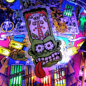 May include: A close-up of a pinball machine with a green zombie-like character sticker. The sticker has a red tongue and a pinball machine design inside. The machine has text that says "FEAR", "FIRE", and "WARNING INSIDE".