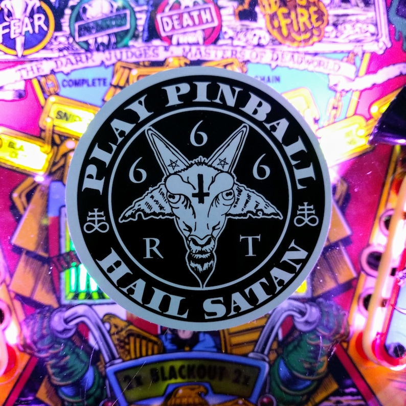 Puede incluir: Una pegatina en blanco y negro con las palabras "Play Pinball Hail Satan" alrededor de una cabeza de cabra con un pentagrama y el n&uacute;mero 666.