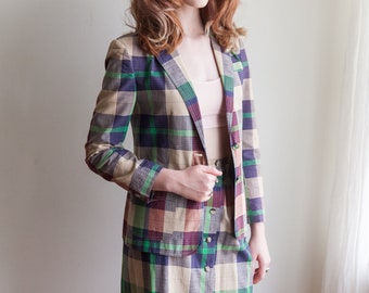 Op de afbeelding: Een geruite rok en blazer set in tinten groen, bruin, beige en paars. De blazer heeft een knoopsluiting en de rok heeft een knoopsluiting aan de voorkant.