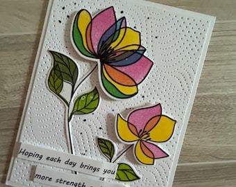 Op de afbeelding: Een witte wenskaart met een gestructureerde, gestippelde achtergrond met twee gelaagde, kleurrijke bloemenillustraties in roze, geel, groen en blauw. Twee witte papieren stroken tonen de tekst: Hoping each day brings you more strength and smiles.