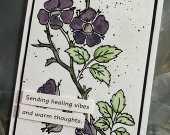 Op de afbeelding: Een rechthoekige wenskaart met een handgetekende illustratie van paarse bloemen en groene bladeren op een witte achtergrond, voorzien van zwarte inktlijnen en spikkels. Twee witte papieren stroken bevatten de tekst Sending healing vibes and warm thoughts.