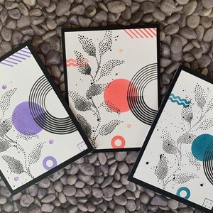 Op de afbeelding: Drie kunstdrukken met zwarte randen, met botanische ontwerpen met gestippelde bladeren en geometrische vormen. Elke print heeft een ander kleuraccent: paars, koraal en blauwgroen. De prints staan op een stenen achtergrond.