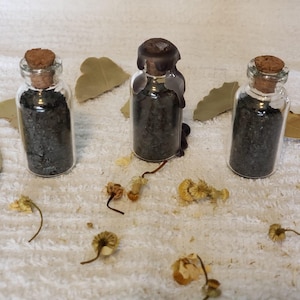 May include: Three small glass bottles filled with dark granules, each sealed with a cork or wax. Dried leaves and small dried flowers are scattered around the bottles on a white surface. The bottles are arranged in a row.