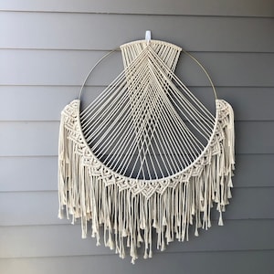 May include: A large, circular macrame wall hanging with a gold metal hoop and a fringe of white cotton yarn. The macrame is woven in a geometric pattern.