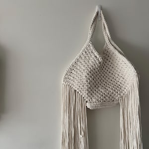 May include: Off-white macrame tote bag with long fringe detail. The bag is made of woven cotton rope and has two handles. The bag is hanging on a white wall. The bag's design is a geometric pattern.