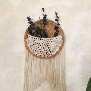 May include: A circular woven wall hanging with a cream macrame pocket and fringe. The natural-toned basket holds dried greenery. The wall decor is suspended from a black hook against a neutral wall.