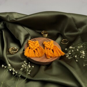May include: A pair of yellow macrame earrings with fringe tassels. The earrings are displayed on a wooden surface with gold rings.