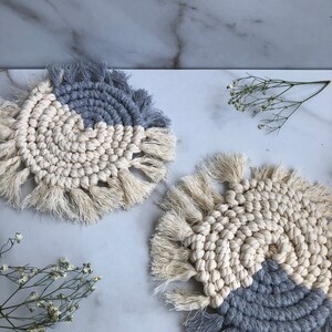 May include: Two handmade crocheted coasters with a white and gray color scheme. The coasters have a fringe border and a circular design.