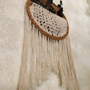 May include: A woven wall hanging with a half-circle design. The top is a brown woven basket shape with a cream-coloured crocheted section. Cream-coloured fringe hangs below. Dark green leaves are placed in the basket.
