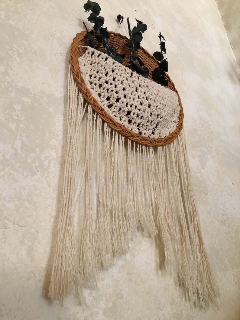 May include: A woven wall hanging with a half-circle design. The top is a brown woven basket shape with a cream-coloured crocheted section. Cream-coloured fringe hangs below. Dark green leaves are placed in the basket.