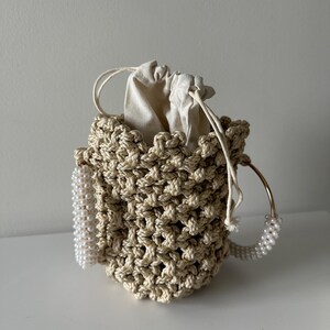 May include: A beige macrame bucket bag with a drawstring closure. The bag features a gold-tone handle and a pearl beaded accent. The interior is lined with a cream-colored fabric.