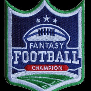 May include: A shield-shaped embroidered patch featuring a blue background with a white football graphic, three stars, and the text FANTASY FOOTBALL in white letters. A red banner below reads CHAMPION, set above a green field with white yard lines.