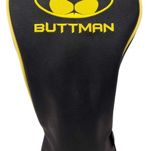 May include: A black golf club head cover with a yellow piped edge. The front features an embroidered yellow oval emblem containing a stylized bat-like shape above the yellow text BUTTMAN in a bold, sans-serif font on a smooth black surface.