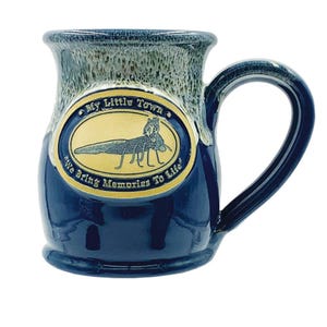 May include: A blue ceramic mug with a gold oval design featuring a dragonfly and the text "My Little Town" and "We Bring Memories To Life".